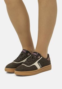 Zapatillas de ante marrón con rayas laterales color crema y acentos en rosa. Presentan un diseño de cordones y suela de goma texturizada. Estilo atlético clásico.