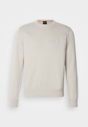 Jasno beżowy sweter wykonany na drutach, z długimi rękawami, okrągłym dekoltem, prążkowanym dołem i mankietami, z małym prostokątnym logo na piersi.