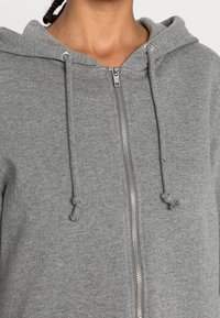 Grijze hoodie met rits gemaakt van zachte stof, voorzien van een capuchon met trekkoord, metalen rits en een subtiele textuur. Geen zichtbare logo's of patronen.