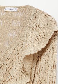 Tissu beige tricoté avec un motif en dentelle ajourée et des fils métalliques subtils, montrant les détails de l'épaule et de l'encolure d'un vêtement.