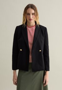 Blazer negro de doble botonadura con botones dorados, llevado sobre una blusa de rayas rojas y blancas. El diseño cuenta con un ajuste entallado y solapas afiladas.