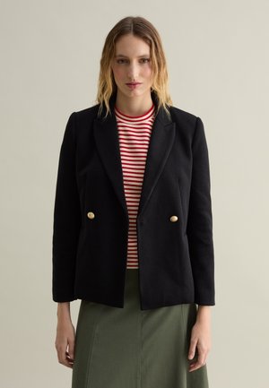 Blazer noir à double boutonnage avec des boutons dorés, porté par-dessus un haut rayé rouge et blanc. Le design présente une coupe ajustée et des revers nets.