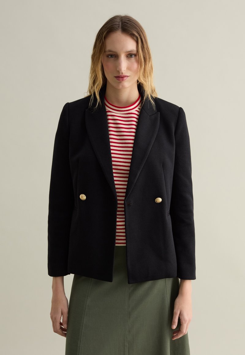 Blazer doppiopetto nero con bottoni dorati, indossato su una maglietta a righe rosse e bianche. Il design presenta una vestibilità sartoriale e rever appuntiti.