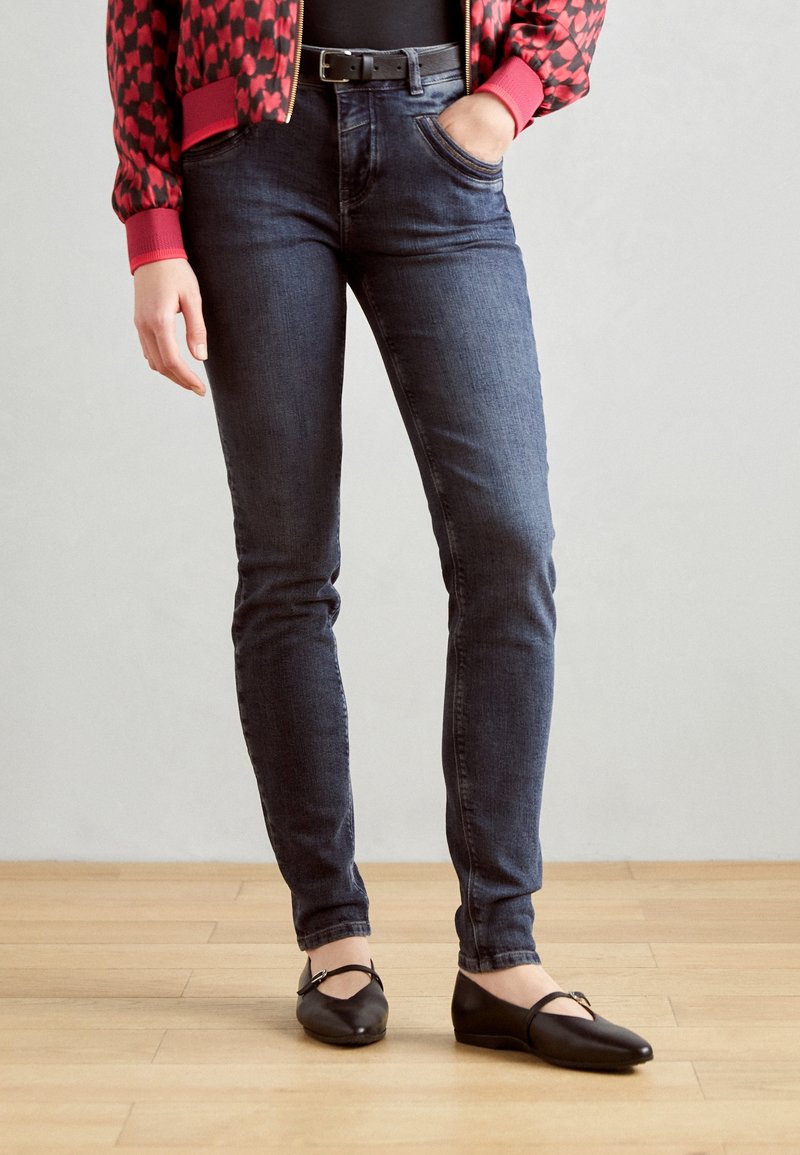Personne portant un jean slim foncé, une ceinture noire, une veste rouge à motifs et des ballerines noires, debout sur un sol en bois. Une main dans la poche.