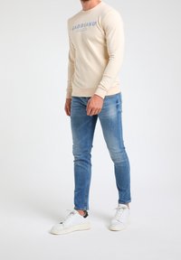 Sudadera beige con el logo "GABBIA" en la parte delantera, combinada con unos vaqueros ajustados de color azul claro y zapatillas blancas con un acento negro en el talón.