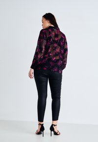 Blouse noire transparente avec un motif floral rose, associée à des leggings noirs brillants et des sandales à talons hauts noires. Vue arrière de la tenue.