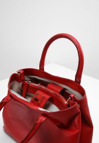 Sac à main en cuir rouge avec texture lisse, compartiment zippé et poignées structurées. L'intérieur présente une doublure claire et un design à plusieurs poches.