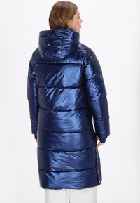 Sötétkék puffázott dzseki fényes felülettel, kapucnival és vízszintes tűzéssel. Oldalsó gombokkal rendelkezik, térd fölé ér és kötött mandzsetta található rajta.