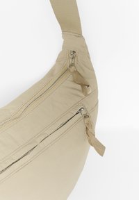 Beige Nylon Umhängetasche mit einer geschwungenen Form, ausgestattet mit einer Reißverschluss-Tasche, strukturiertem Tragegurt und metallischen Reißverschluss-Beschlägen.
