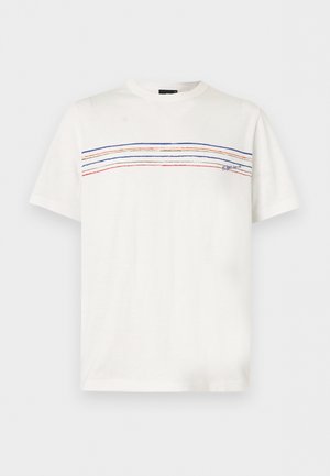 Homme portant un t-shirt blanc à manches courtes avec des rayures colorées, un jean bleu coupe décontractée et des mocassins beiges, debout les mains derrière le dos.