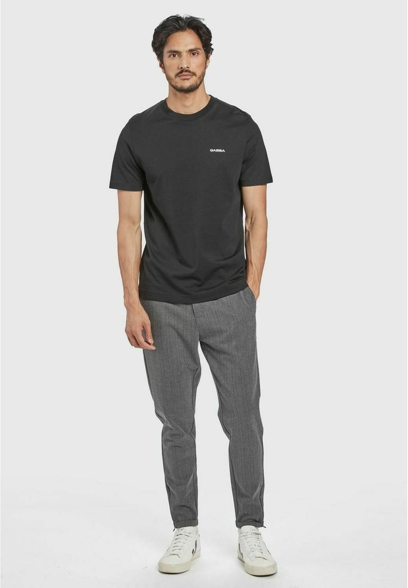 Schwarzes Baumwoll-T-Shirt mit Rundhalsausschnitt und kleinem weißen Logo, kombiniert mit grauen gemusterten Hosen und weißen Sneakers.