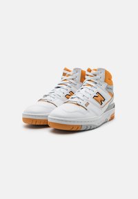 Białe sneakersy typu high-top z pomarańczowymi akcentami, teksturowaną skórą, wyściełanym kołnierzem oraz szarym śródpodłożem z żebrowaną gumową podeszwą.