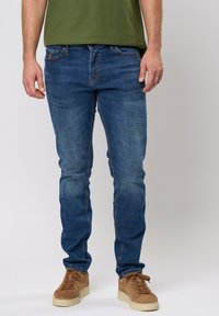 Jean en denim bleu moyen avec une coupe slim, présentant un léger délavage, cinq poches et une patch en cuir marron sur la taille arrière.