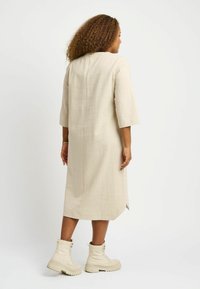 Knehöydylle yltävä beige pellavamekko, jossa on kolmanneksen hihat, takasauman yksityiskohtia ja sivuhalkioita, yhdistettynä kermaisiin nauhakuulotsinukkeihin.