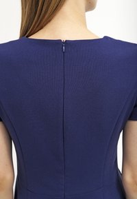 Bakifrån av en person som bär en figurnära marinblå klänning med en kort dragkedja på övre delen av ryggen och korta ärmar.