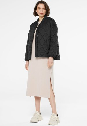 Frau trägt eine schwarze gesteppte Jacke über einem beigen Midikleid mit seitlichem Schlitz und weißen Sneakern, steht vor einem einfarbigen weißen Hintergrund.