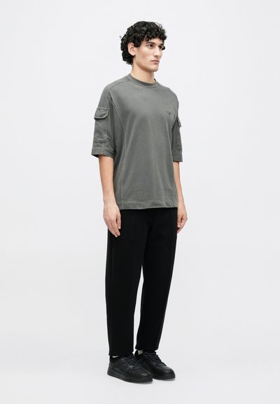 Šedé oversized tričko s krátkými rukávy a logem na hrudi, kombinované s černými zúženými kalhotami a černými teniskami, na jednobarevném pozadí.