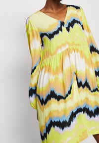 Vestido con estampado floral en amarillo, verde y negro. Presenta un escote en V, mangas largas abullonadas y un diseño en capas con una tela ligera.