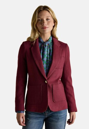 Blonde Frau mit einem burgunderroten Blazer über einer blau-grün gemusterten Bluse und blauen Jeans, stehend vor einem weißen Hintergrund.