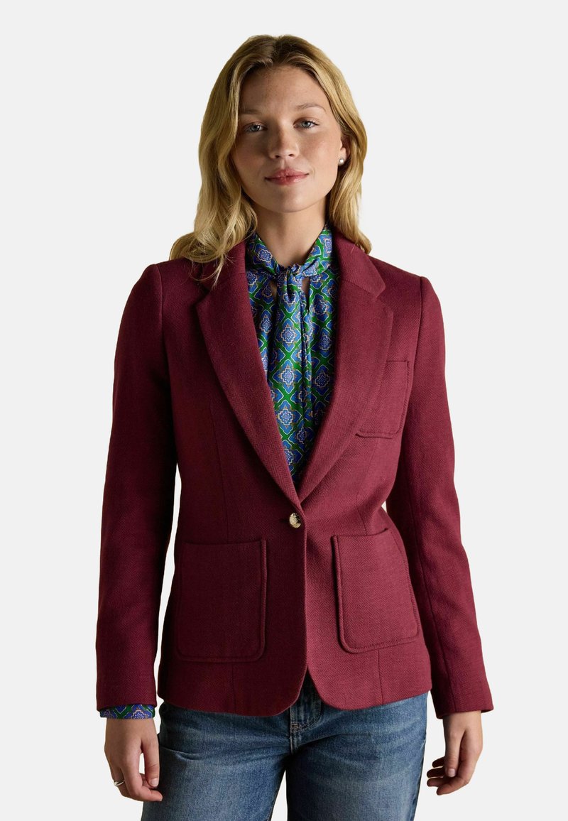 Blond naine, kes kannab burgundi värvi bleiserit, sinise ja rohelise mustriga pluusi ning siniseid teksaseid, seistes valge tausta ees.