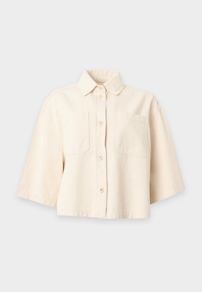 Chemise beige crème à manches courtes avec col et deux poches poitrine, présentée sur un fond clair uni.