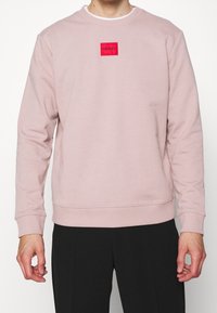 Sudadera de color rosa claro con cuello redondo, puños acanalados y un parche cuadrado de logo rojo en el pecho, fabricada en suave tela de algodón.