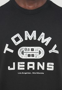 Μαύρο κοντομάνικο μπλουζάκι με έντονο λευκό γραφικό κείμενο «TOMMY JEANS 1985» και σύμβολο αλτήρα, με υφασμάτινη υφή.