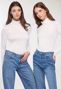 Dwie modelki ubrane w białe swetry z wysokim dekoltem i jasnoniebieskie dżinsy z wysokim stanem. Swetry mają dopasowany krój, a dżinsy posiadają klasyczny pięciokomorowy fason.