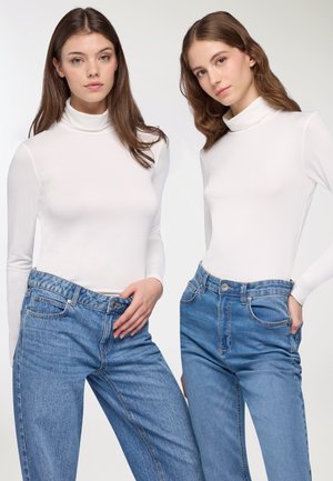 Due modelle indossano maglioni bianchi a collo alto e jeans in denim azzurri a vita alta. I maglioni sono attillati e i jeans hanno un classico design a cinque tasche.