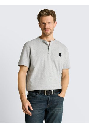 Mężczyzna z lekkim zarostem, ubrany w jasnoszarą koszulkę henley z krótkim rękawem i ciemnoniebieskie dżinsy, stoi z jedną ręką w kieszeni na jednolitym tle.