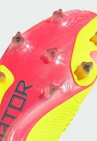 Semelle de chaussure de football avec une surface rose vif et des accents jaunes. Présente plusieurs crampons transparents pour une traction optimale et des motifs texturés pour une bonne adhérence.