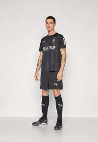 Svart fotbollströja med nätstruktur, prydd med vita logotyper och detaljer. Kombinerad med svarta shorts, knäla långa svarta strumpor och fotbollsskor.