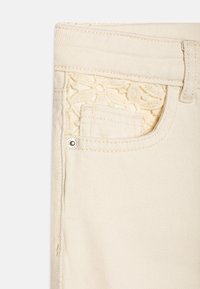 Kremfargede denimjeans med et blondermønster inni frontlommen og en metallknappdetalj på linningen.