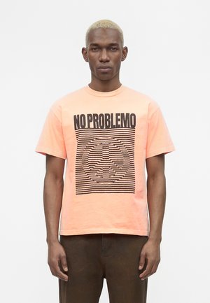 Homme aux cheveux courts décolorés portant un t-shirt pêche avec le texte « NO PROBLEMO » et un symbole de paix composé de lignes noires horizontales.