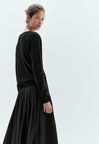 Maglione nero a maglia con maniche lunghe e polsini aderenti, abbinato a una gonna a pieghe nera. Texture liscia, design minimalista su uno sfondo chiaro.