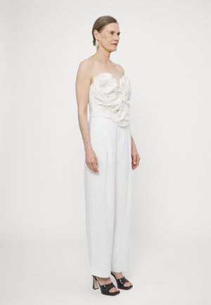 Hvid strapless jumpsuit med store stofblomster på overdelen, bredbenet design og kontrasterende sorte højhælede sandaler.