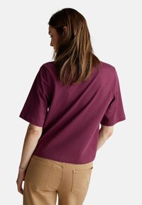 T-shirt in cotone borgogna con maniche corte e vestibilità morbida; indossato con pantaloni beige, con vista posteriore e superficie liscia.