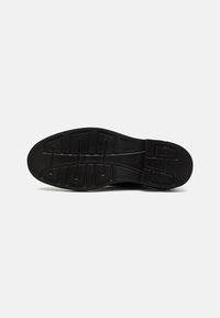 Semelle de chaussure noire avec des motifs de relief géométriques profonds et un petit code embossé sur un fond clair uni.