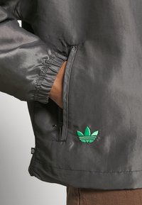 Person wearing black jacket with hand in zipped pocket and green Adidas trefoil logo on front near waist.