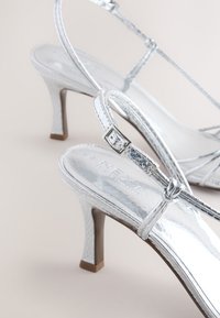 Ezüst színű, magas sarkú szandálok fényes felülettel, vékony pántokkal és kis csattal. Texturált sarokkal és minimalista dizájnnal rendelkeznek.