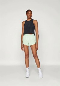 Nő fekete ujjatlan toppban, világoszöld sportnadrágban, fehér zoknikban és fehér sportcipőkben áll egy sima világos háttér előtt.