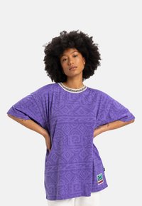 Fialové oversized tričko s zložitými geometrickými vzormi, s kontrastným golierom a logom umiestneným na spodnej strane.