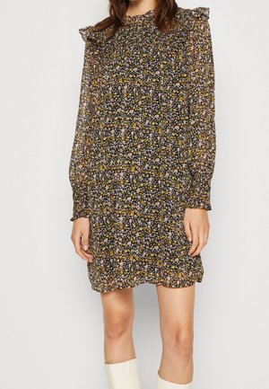 Kobieta w mini sukience z długim rękawem w kwiatowy wzór, z falbankami na ramionach i białych kozakach za kolano.