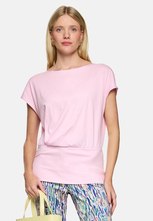 Frau trägt ein hellrosa kurzärmeliges Oberteil und bunt gemusterte Hose, hält eine blassgelbe Handtasche vor weißem Hintergrund.