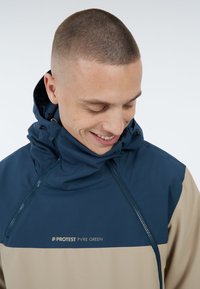 Zweifarbige Pulloverjacke in Marineblau und Beige, mit hohem Kragen, verstellbarem Kapuze, Frontreißverschluss und dem "PROTEST PVRE GREEN"-Logo.