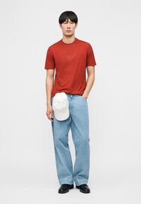 Άνδρας που φοράει κόκκινο μπλουζάκι, ανοιχτό μπλε παντελόνι με φαρδιά μπατζάκια, μαύρα παπούτσια, κρατώντας λευκό καπέλο, στέκεται μπροστά σε απλό λευκό φόντο.