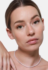 Przezroczysta choker z koralikami z małymi, gładkimi, półprzezroczystymi koralikami. Naturalny kolor z błyszczącym wykończeniem. Modelka prezentuje ją na neutralnym tle.