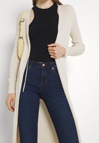 Lång ribbad cardigan i beige över en svart ärmlös topp, tillsammans med mörkblå jeans; inkluderar en liten gul väska med axelrem.