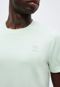 Άνδρας που φοράει ανοιχτό πράσινο υφασμάτινο κοντομάνικο πουκάμισο με μικρό ανάγλυφο λογότυπο "F" στο αριστερό μέρος του στήθους.