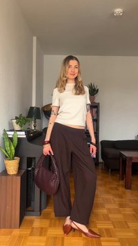 Γυναίκα με μακριά ξανθά μαλλιά στέκεται μέσα σε κλειστό χώρο, φοράει λευκό τοπ, καφέ παντελόνια με φαρδιά μπατζάκια και καφέ μυτερά φλατ παπούτσια, κρατώντας ένα πλεκτό καφέ τσάντα.